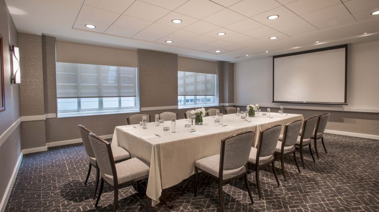 A conference room with a rectangular table, chairs, a projector screen, and a lit ceiling. The table is set with glasses and decorative items.