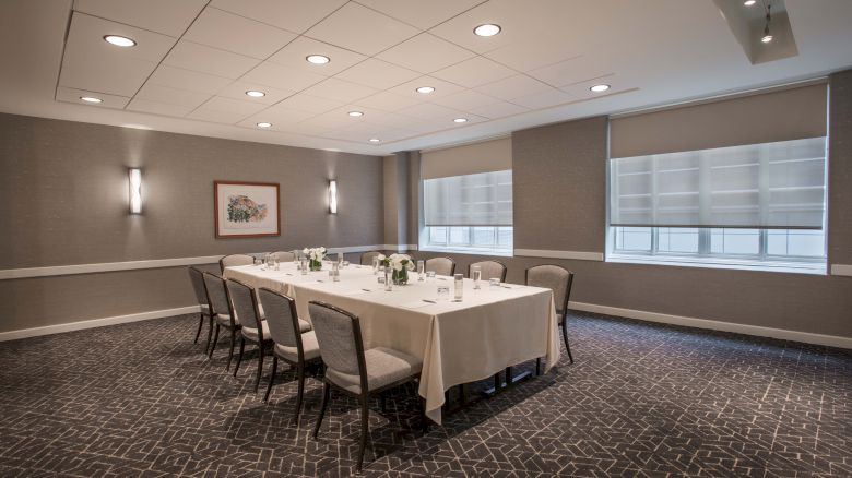 A conference room with a U-shaped table setup, surrounded by chairs, with lighting and blinds drawn.