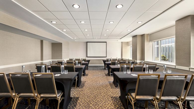 A well-lit conference room with rows of tables and chairs facing a screen, ready for a meeting or presentation.