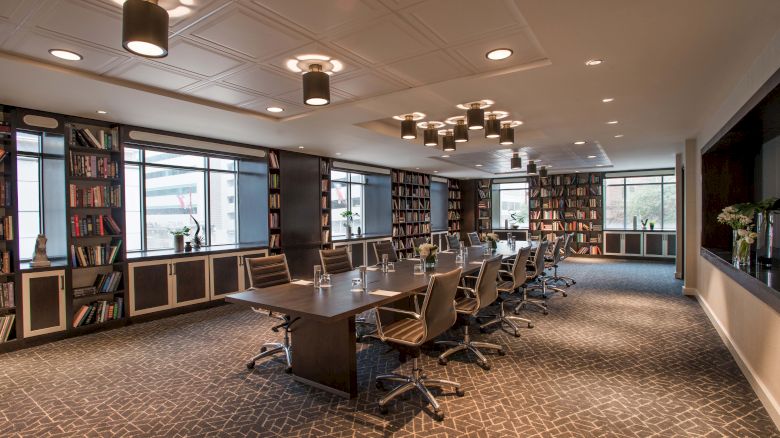 A modern conference room with a long table, chairs, bookshelves, and large windows, featuring overhead lighting for a professional setting.