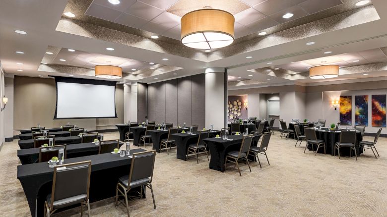 A conference room set up with tables and chairs facing a large screen, featuring modern lighting and abstract wall art on the sides.