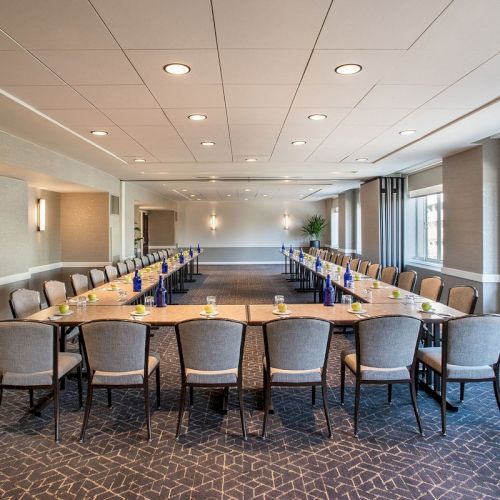 The image shows a modern conference room with a large U-shaped table, chairs, and water bottles, featuring ample natural light from windows.