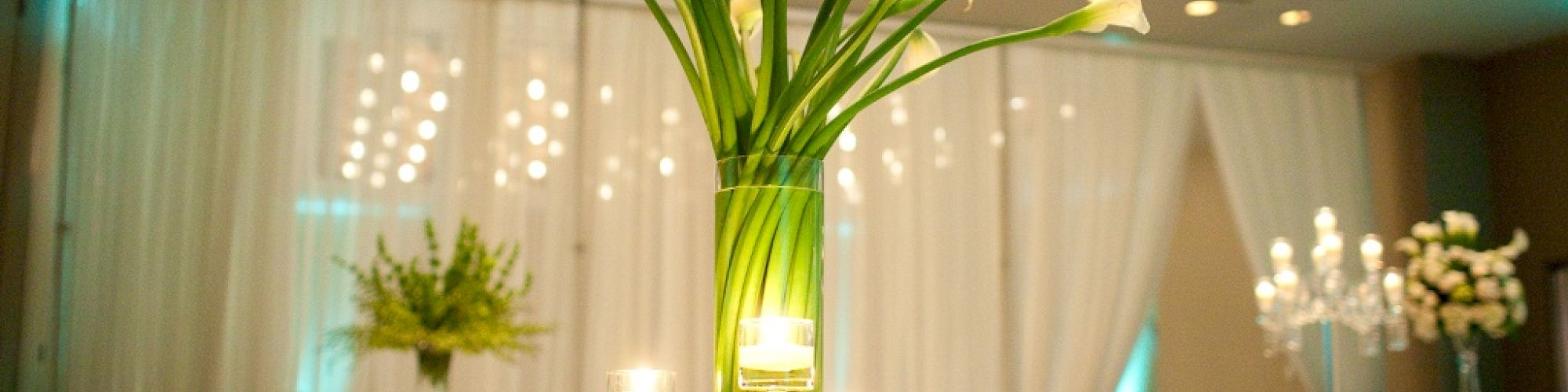 A beautifully set table at an elegant event with white flowers in a tall vase, candles, and a softly lit ceiling.