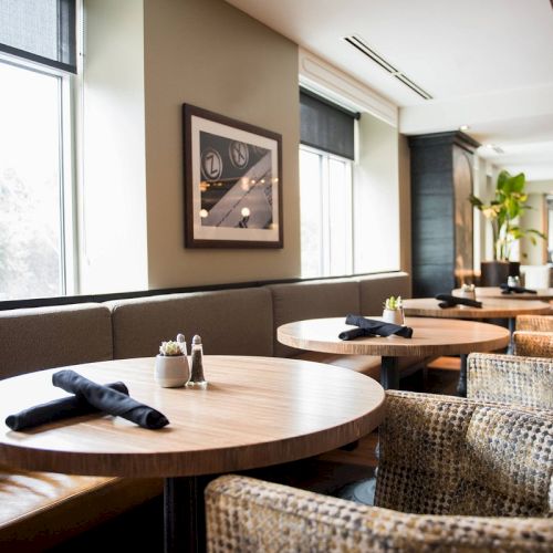 A cozy restaurant interior with round tables, chairs, and large windows. Black napkins are placed on the tables, creating an inviting atmosphere.