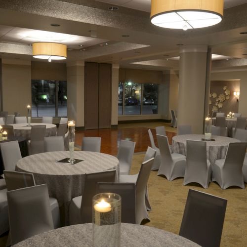 The image shows an elegantly decorated event space with round tables, gray chairs, and candles, ready for a gathering or function.