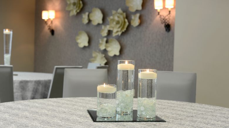 A table with three glass candle holders, floral wall decor, and soft wall lighting in a modern room setting.