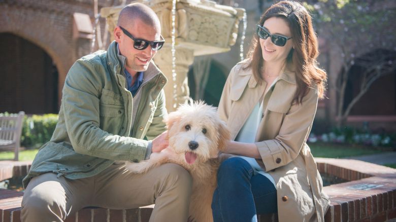 Two people wearing sunglasses sit by a fountain, smiling and petting a fluffy dog.