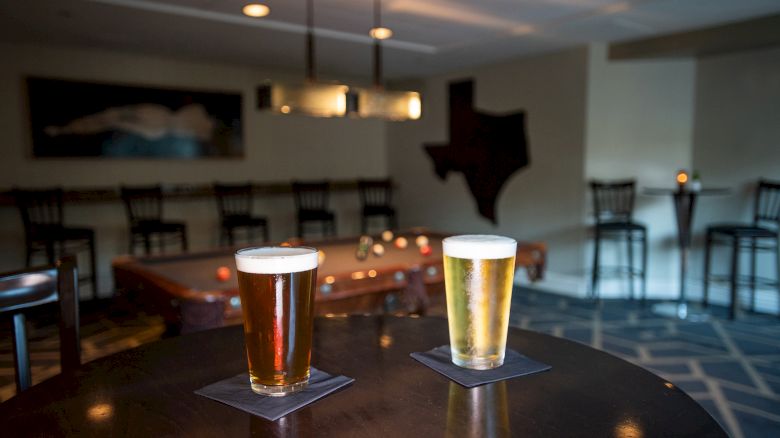 Two glasses of beer on a dark table in a dimly lit bar with Texas-shaped wall decor and seating in the background.