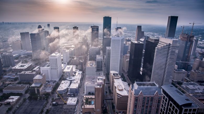 Aerial view of a cityscape with tall skyscrapers, clouds drifting between buildings, and a hazy sunlit sky.