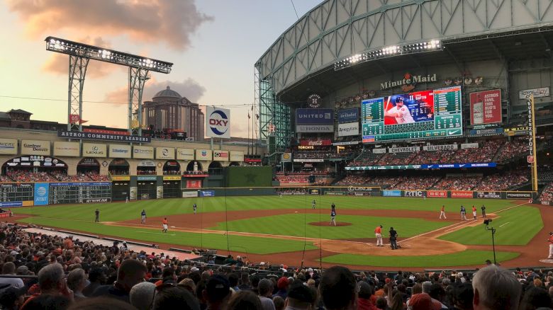 A baseball game is in progress at a large stadium, filled with spectators under a clouded sky as evening approaches.