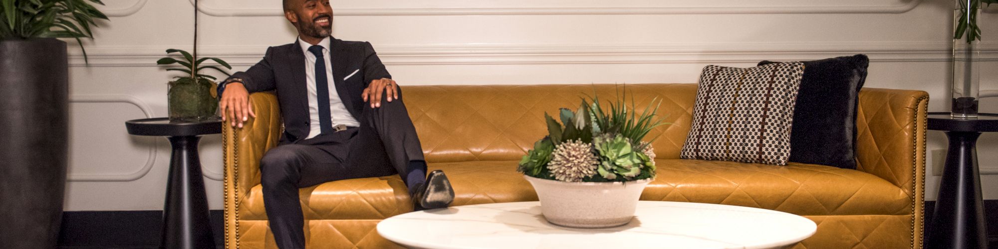 A person in a suit sits on a yellow sofa with a modern coffee table and plants, under a decorative mirror in an elegant interior setting.