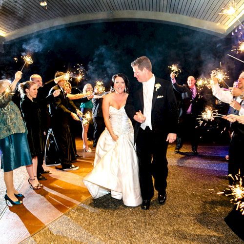 A wedding couple walks through a line of guests holding sparklers in a celebratory atmosphere indoors.