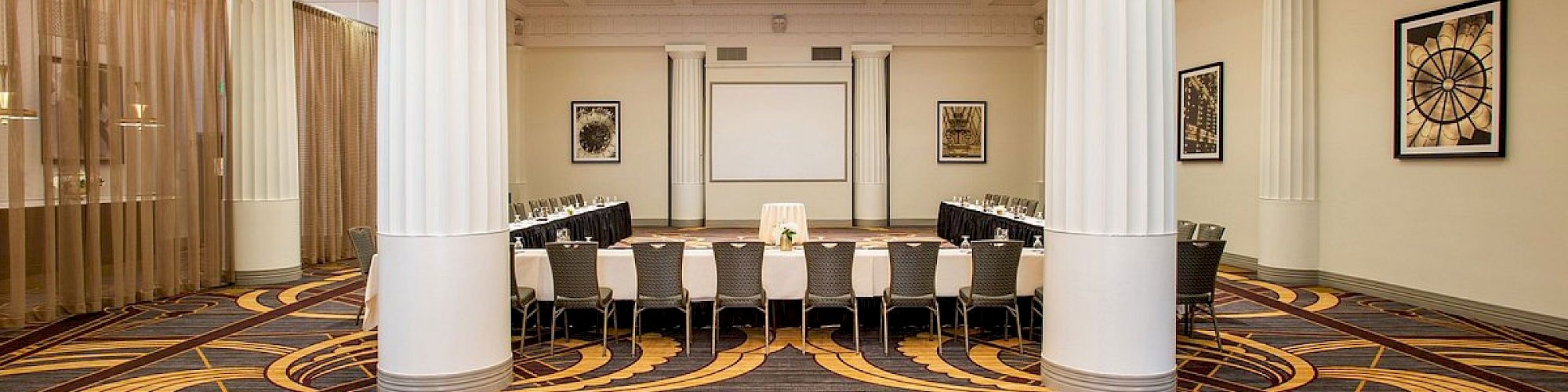 A conference room with a long table surrounded by chairs, ornate carpet, large columns, and a ceiling with decorative lighting fixtures.