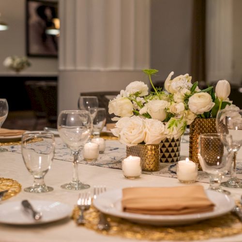 Elegant table setting with white floral centerpiece, wine glasses, golden accents, and neatly arranged silverware on a cream-colored tablecloth.