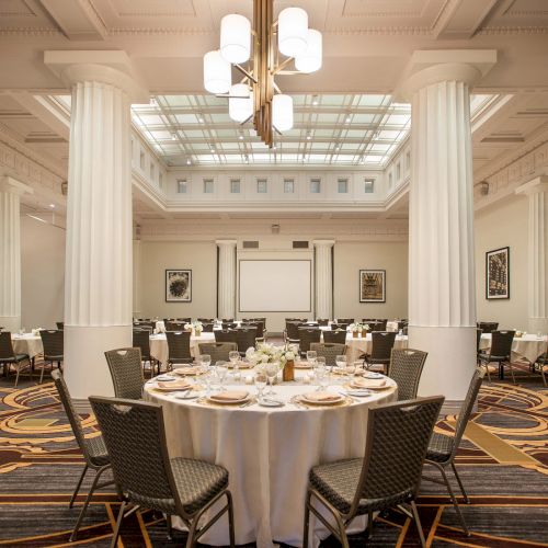 The image shows an elegant banquet hall with round tables, set with plates and cutlery, surrounded by chairs and lit by chandeliers.