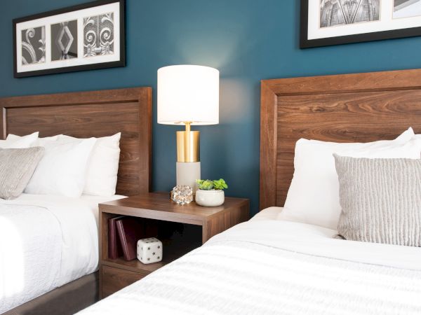 The image shows two beds with wooden headboards, a shared nightstand with a lamp and decor, against a blue wall, and framed artwork.