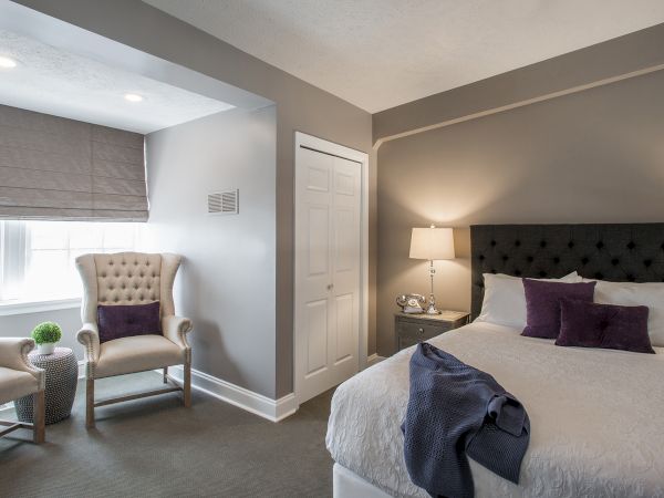 A cozy bedroom with a bed, two chairs by a window, a bedside table with a lamp, and decorative pillows and a throw blanket.