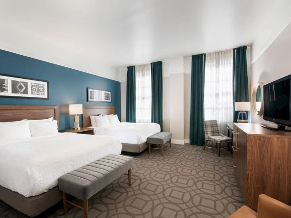 The image shows a hotel room with two beds, teal curtains, a TV on a wooden stand, lamps, and a geometric-patterned carpet.