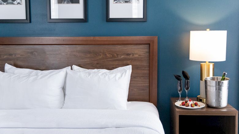 A cozy bedroom with a wooden headboard, three pillows, side table, lamp, champagne in an ice bucket, and a plate of berries.