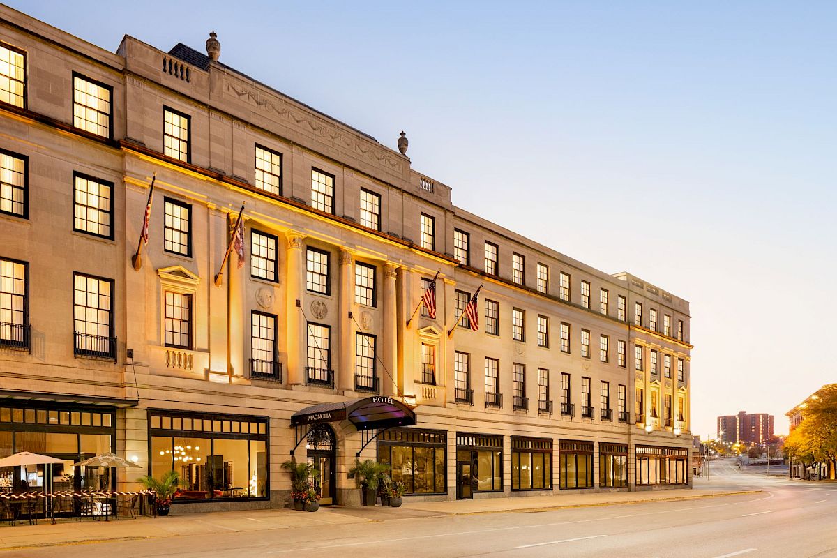 The image shows a grand, multi-story building with flags, large windows, and a welcoming entrance, likely a historic hotel or venue.