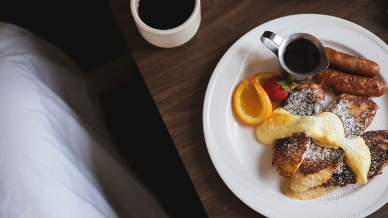 A breakfast plate with French toast, sausages, orange slice, strawberry, powdered sugar, and syrup; accompanied by a cup of coffee.