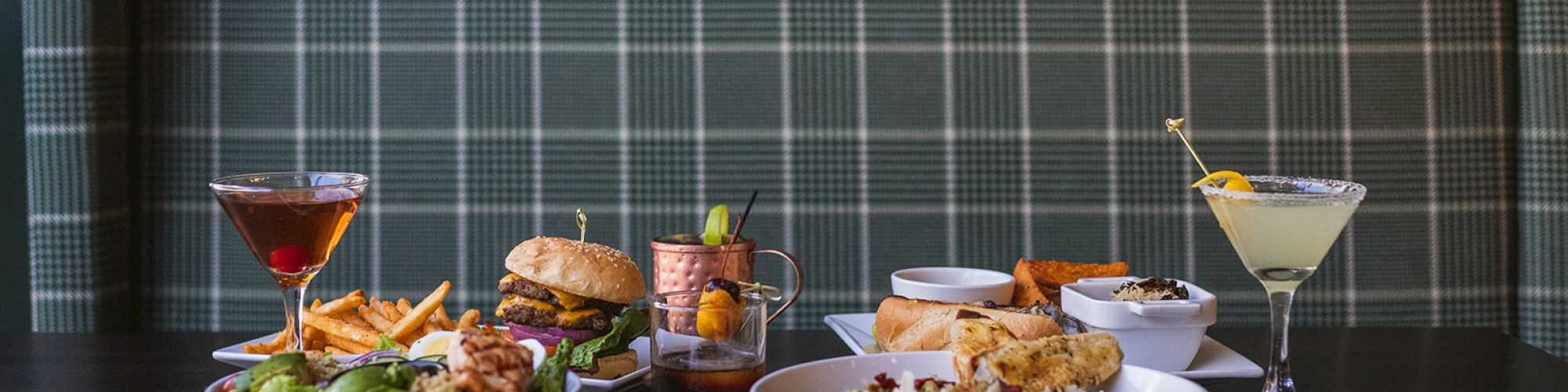 A table with various dishes, including burgers and salad, and drinks like cocktails and soda, set against a plaid upholstered booth.