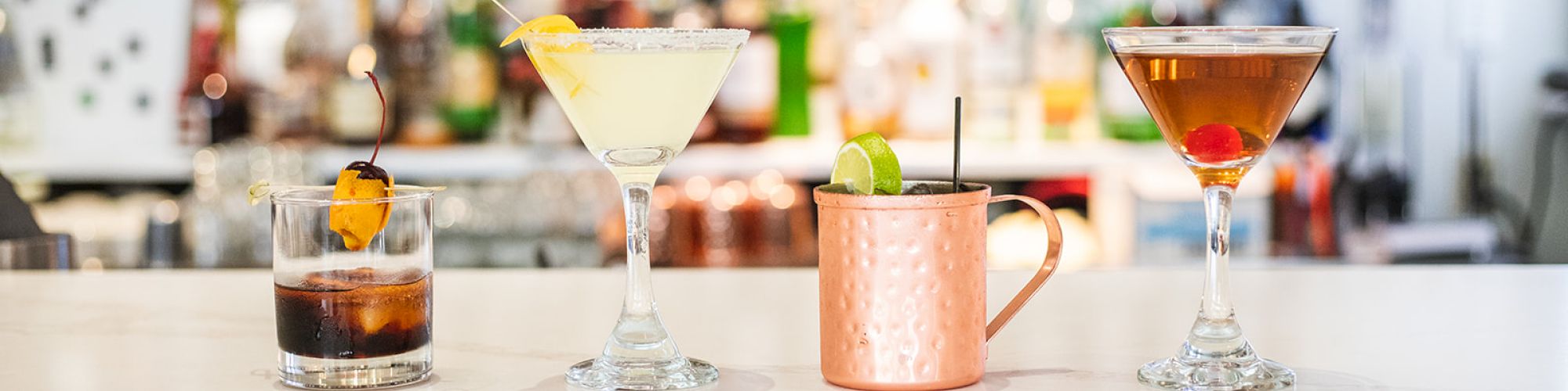 Four cocktails sit on a bar counter: an Old Fashioned, a Margarita, a Moscow Mule, and a Manhattan, with bottles in the background.