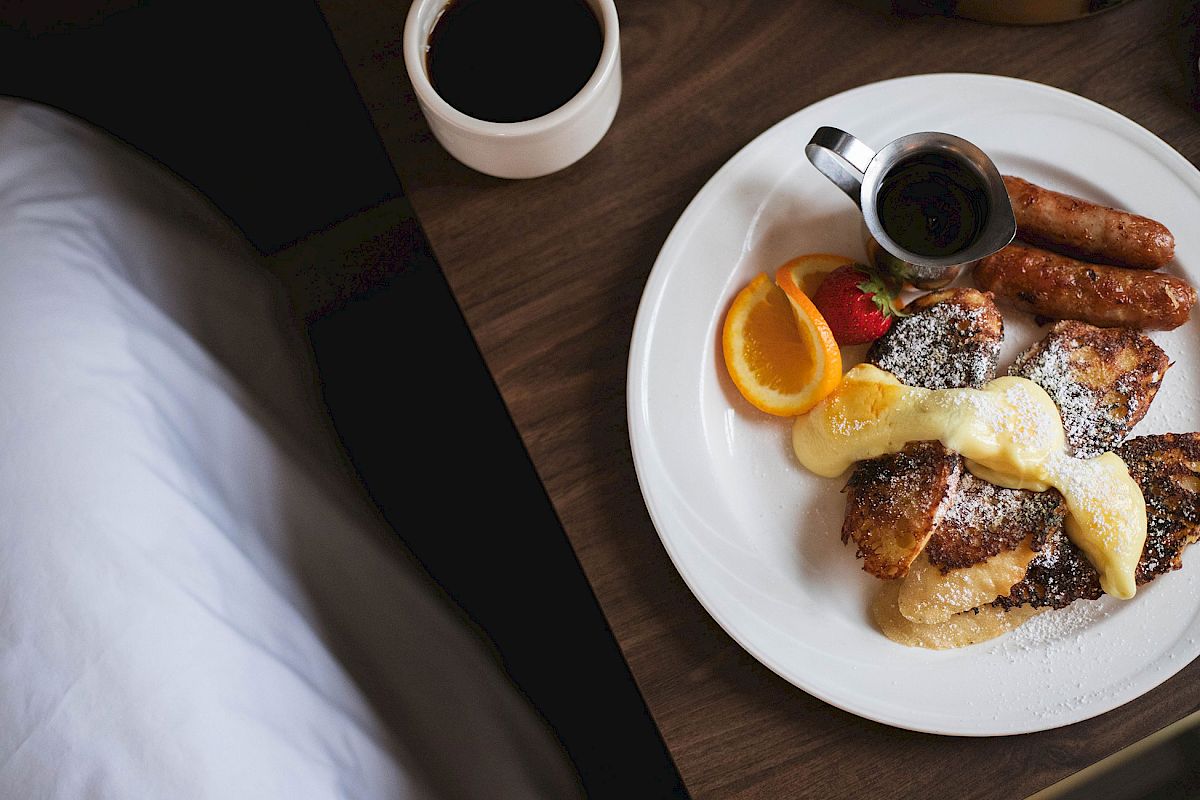 A breakfast plate with sausages, French toast, syrup, powdered sugar, and fruit, next to a cup of coffee on a wooden table.