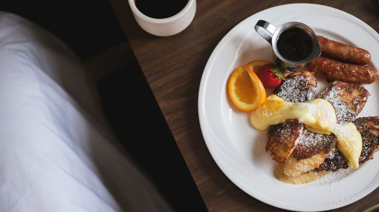 A breakfast plate with sausages, French toast, syrup, powdered sugar, and fruit, next to a cup of coffee on a wooden table.