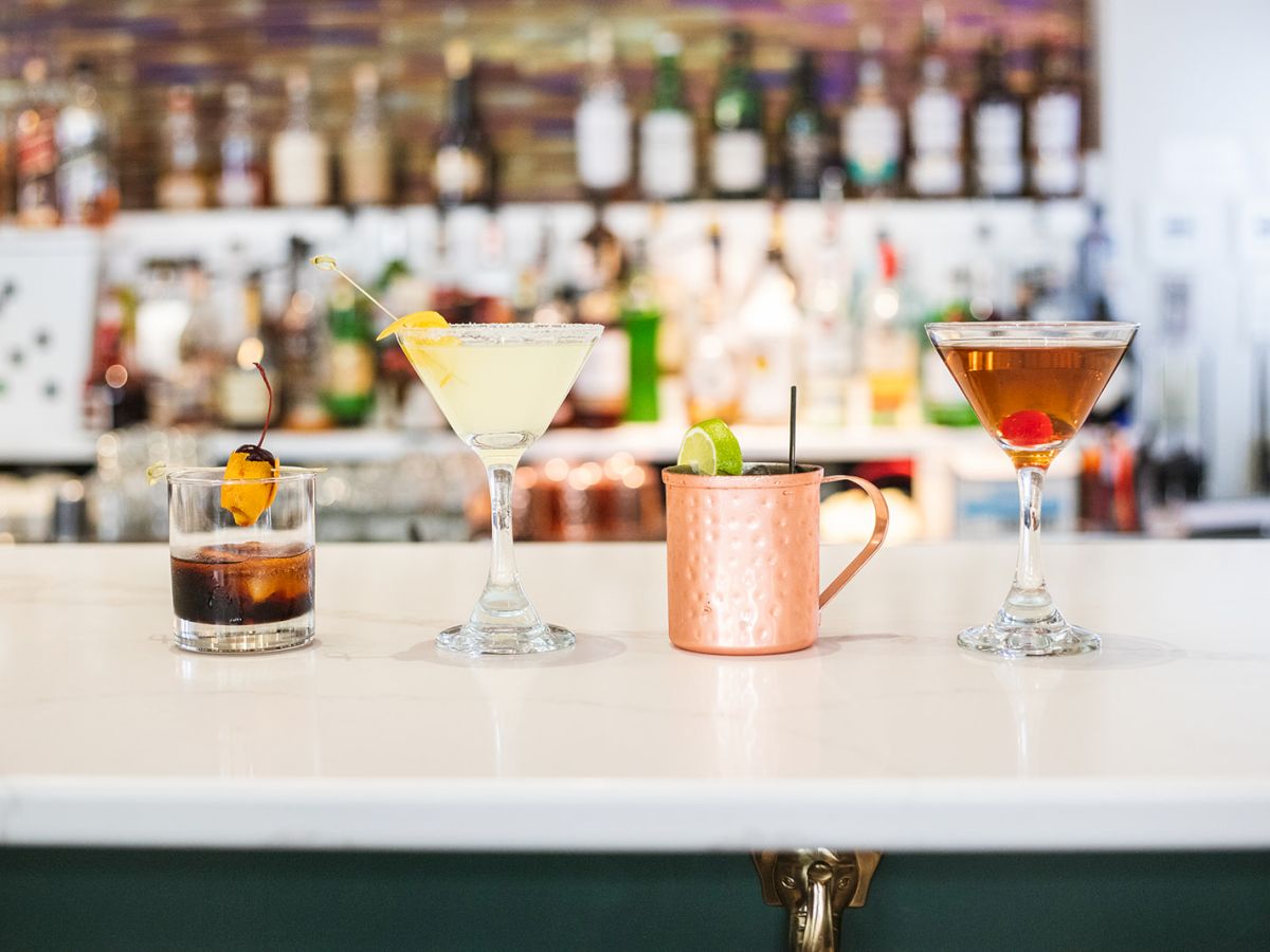 Four cocktails are arranged on a bar counter, each in different glassware, with a blurred bar background.