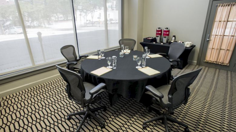 A conference room with a round table, chairs, notepads, glasses, and a refreshments station with coffee and condiments in the corner.