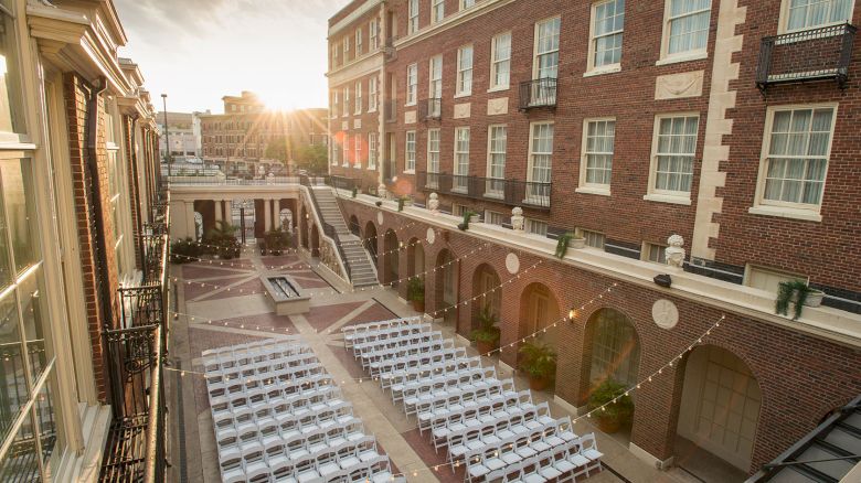 The image shows a courtyard set up for an event with rows of white chairs, surrounded by brick buildings, and illuminated by hanging lights.