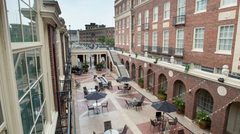 An outdoor courtyard with seating arrangements, surrounded by brick buildings, featuring tables with umbrellas and archways.