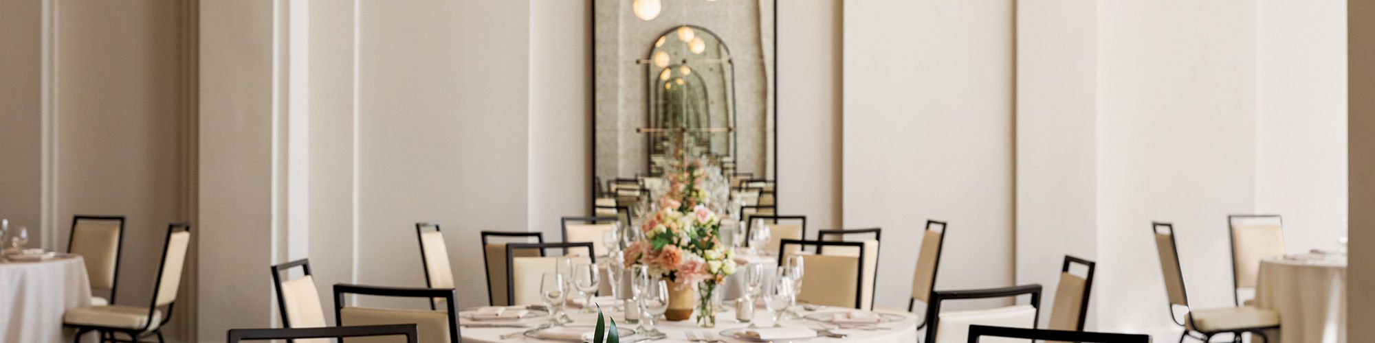 Elegant banquet hall with round tables, floral centerpieces, and set dinnerware. Large mirror and soft lighting enhance the ambiance.