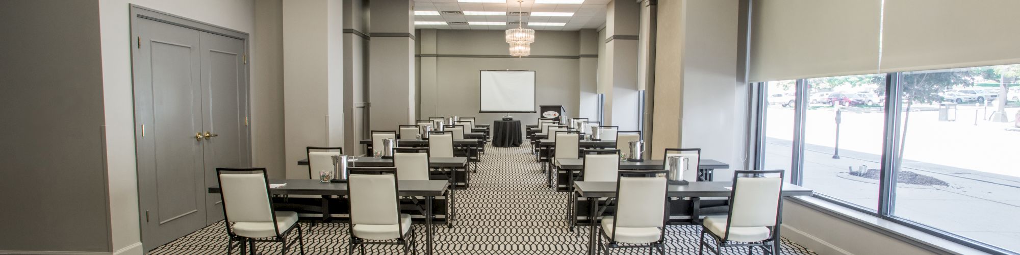 The image shows a modern conference room with rows of tables and chairs, a large window, patterned carpet, and a ceiling chandelier.