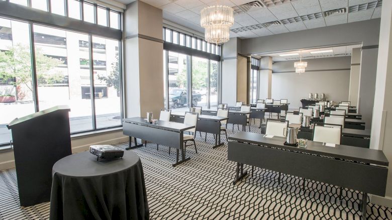 A conference room with arranged tables and chairs, large windows, chandeliers, and a podium with a projector.