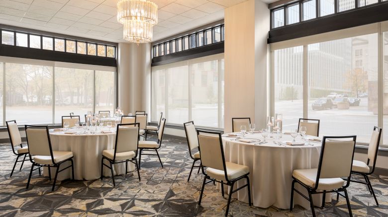 The image shows a banquet room with round tables set for an event, featuring chairs, place settings, and a chandelier, all in a bright space.