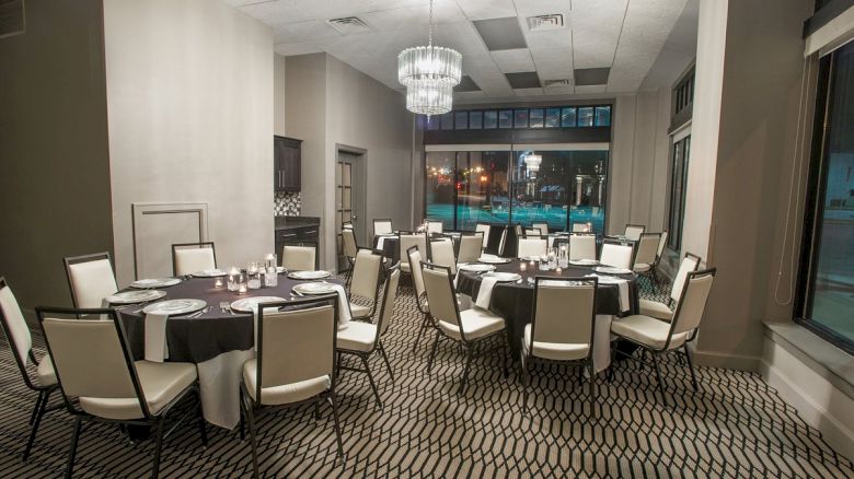 The image shows a dining room with round tables and white chairs, elegantly set up with glassware under a modern chandelier.