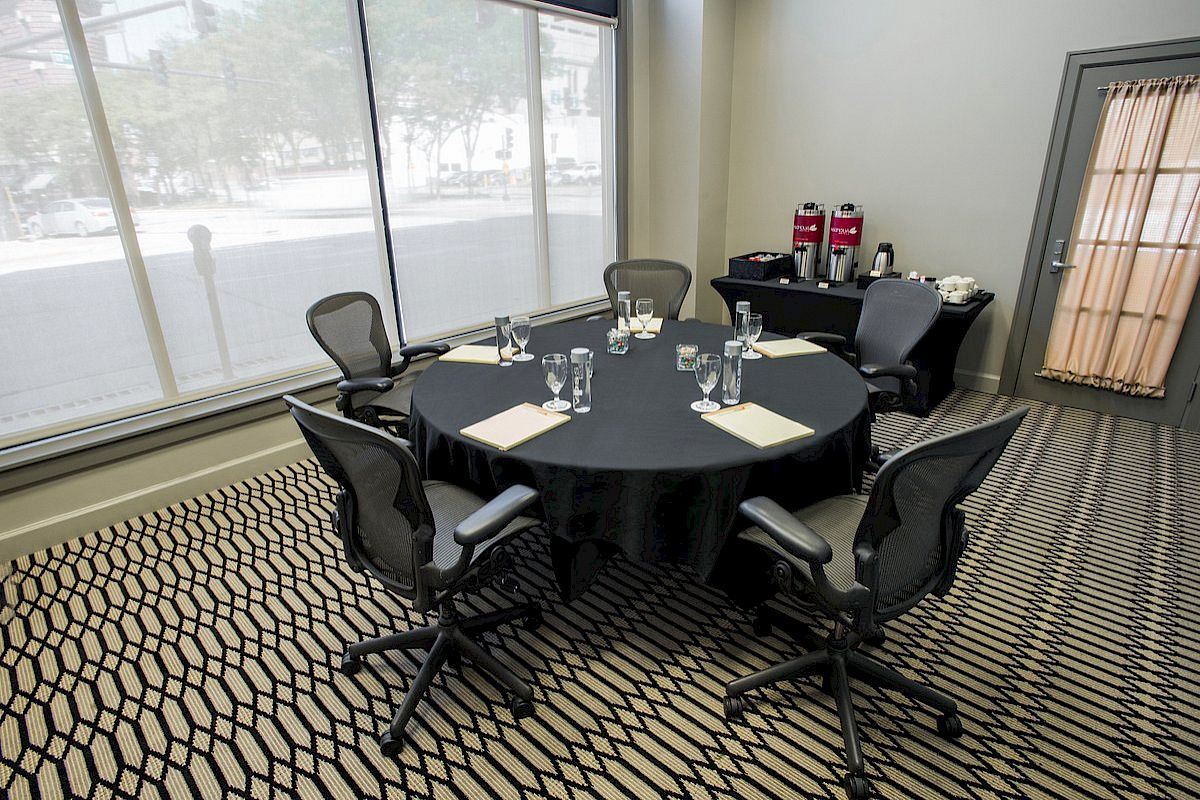 A meeting room with a round table, chairs, glasses, and notepads. There are drinks and a coffee setup on a table by the door.