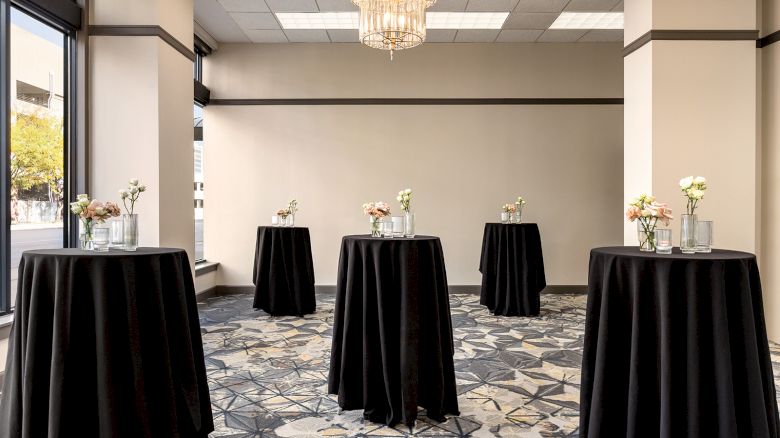 The image shows an elegant room setup with tall tables covered in black cloth, each adorned with small floral arrangements.