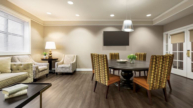 The image shows a modern living room with a round dining table, chairs, a sofa, armchairs, a lamp, and a wall-mounted TV.