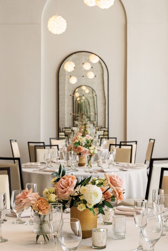 The image shows an elegantly set dining room with round tables, floral centerpieces, modern chairs, and ambient lighting fixtures.