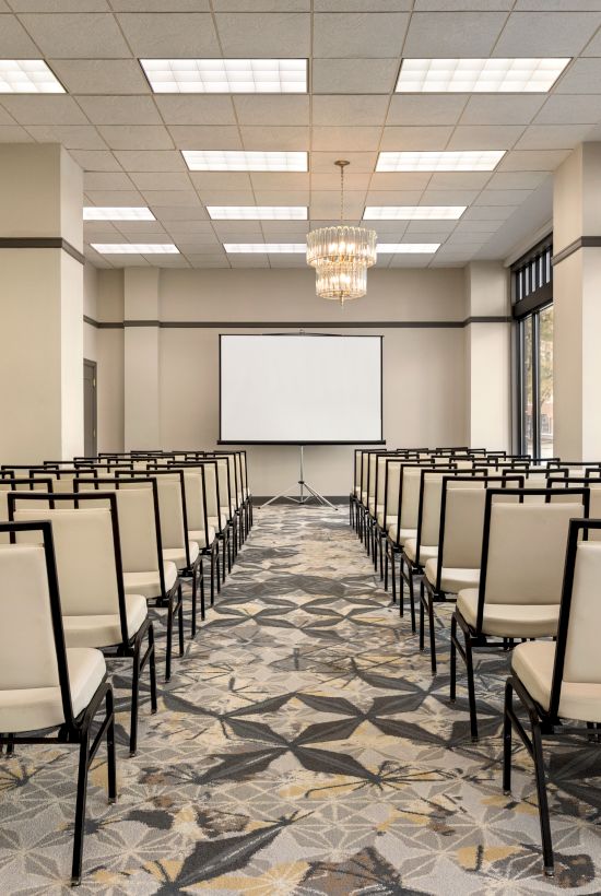 A conference room with rows of beige chairs facing a white screen, under a ceiling with light fixtures, and large, bright windows.