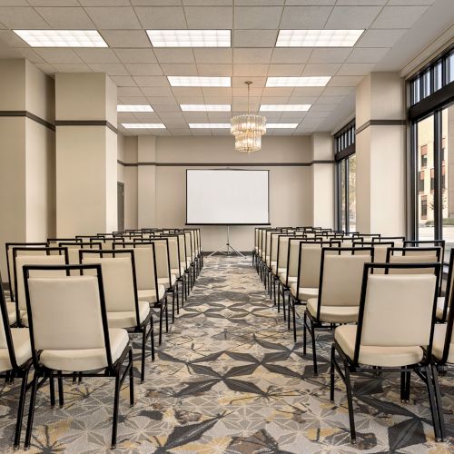 The image shows a conference room with neatly arranged chairs facing a screen at the front, with large windows on the side.