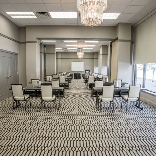 The image shows an empty conference room with rows of tables and chairs, a patterned carpet, and chandeliers.