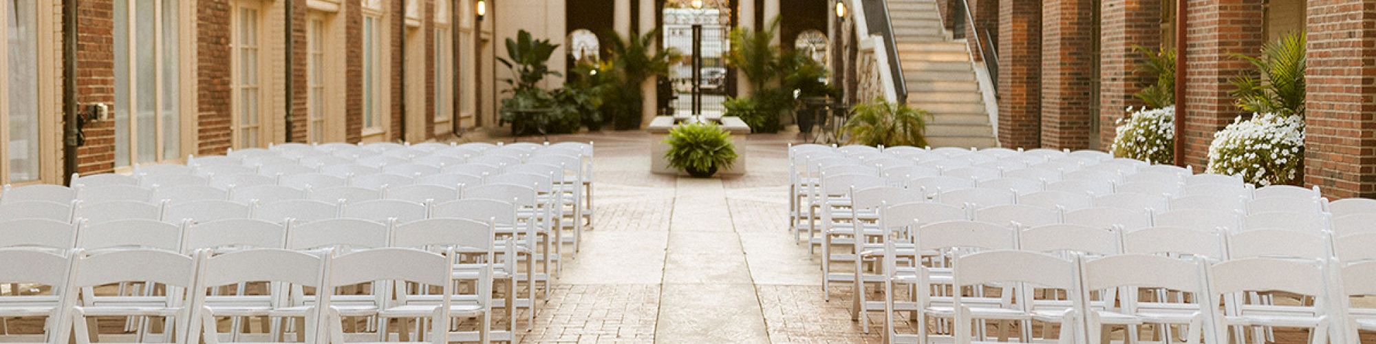 An outdoor event space with rows of white chairs, brick walls, and a stairway, likely set up for a wedding or gathering.