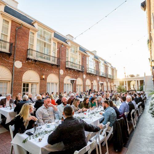 People dining outdoors at long tables between brick buildings, under string lights, in a courtyard setting.