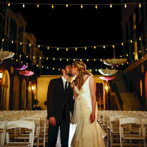 A couple kisses in an outdoor wedding setting with decorative lights and hanging umbrellas. Rows of chairs fill the foreground of the romantic scene.