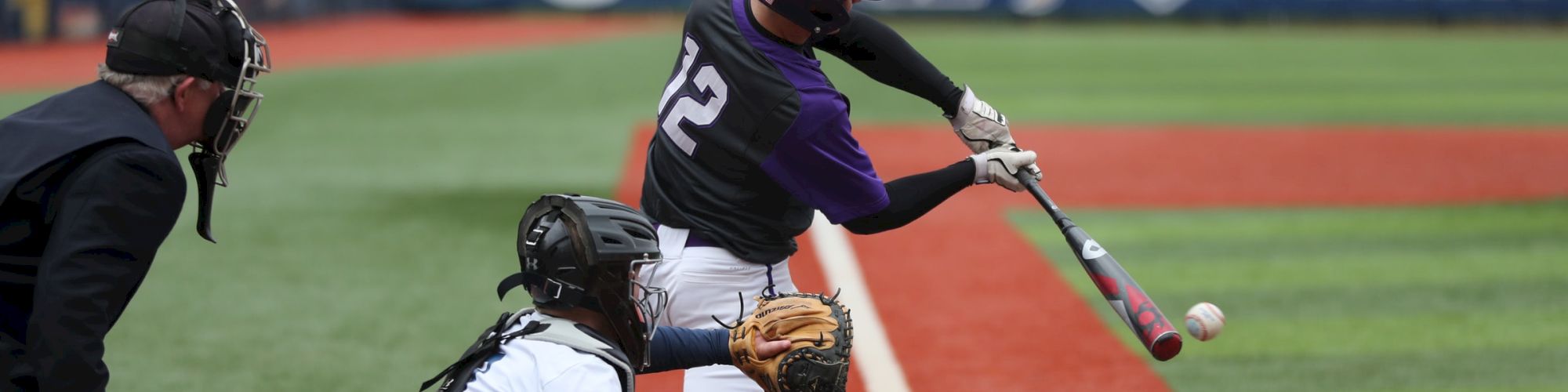 A baseball player in a purple jersey is swinging a bat at a ball, with a catcher and umpire nearby, on a field.