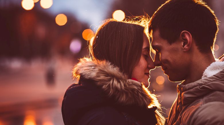 A couple in winter clothes stand closely together, gently touching foreheads, with a blurred, warmly lit urban background.